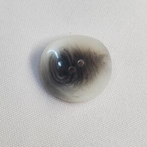 May include: Oval button with a swirling pattern of black and grey within a translucent, off-white material. The button has two small holes and is set against a white fabric background.
