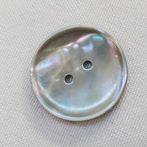 May include: A large, iridescent, oval-shaped button with two holes. The button has a pearlescent sheen with hints of blue, green, and pink. The button is set against a light beige fabric background.