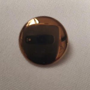 May include: A circular, polished rose gold-toned metal button with a highly reflective, mirror-like surface. The smooth, convex face captures a dark reflection of the surrounding environment, including a small rectangular camera lens shape.