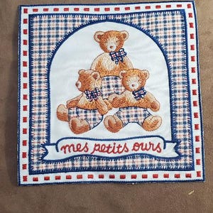 Peut inclure: Carré brodé avec trois ours en peluche portant des pantalons à carreaux bleus et des nœuds papillon. Les ours sont placés sur un fond blanc avec une bordure à carreaux bleus et rouges. Le texte "mes petits ours" est en dessous.