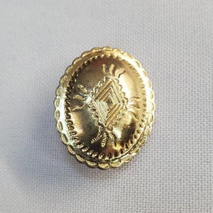 Könnte beinhalten: Ein goldfarbener, runder Knopf mit geprägtem geometrischem Design. Der Knopf hat einen gewellten Rand und eine zentrale Raute, die von radialen Linien umgeben ist. Wahrscheinlich für dekorative Zwecke verwendet.