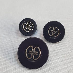 Puede incluir: Tres botones de color púrpura oscuro con un diseño decorativo plateado. Los botones son redondos y ovalados, con un acabado brillante. Los botones están sobre una superficie blanca texturizada.