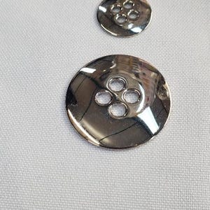 Pode incluir: Dois botões prateados brilhantes com quatro furos cada. O botão maior é oval e o menor é redondo. Ambos os botões refletem a luz, sobre um fundo de tecido branco.
