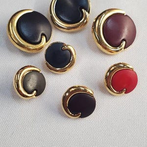 May include: Seven round buttons with a gold-coloured trim. The buttons have a curved design and come in various colours, including black, burgundy, grey, and red. The buttons are arranged on a white surface.