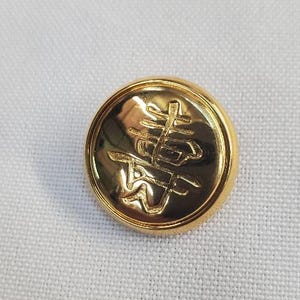 May include: A circular, gold-coloured button featuring an embossed Chinese character design. The button is set against a white, textured background. This button is likely for clothing or craft projects.