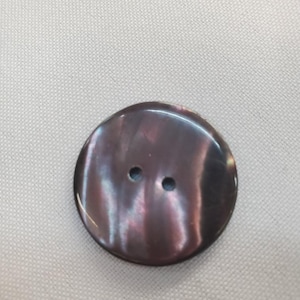 May include: A large, round button with a lustrous, iridescent surface. The button is a dark, purplish-brown colour with two small holes for sewing. The button is set against a neutral, textured background.