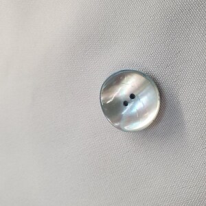 May include: A round, iridescent button with two holes, set against a light gray fabric background. The button has a pearlescent sheen, reflecting various colors. This button is likely for clothing or craft projects.