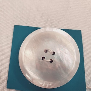 Puede incluir: Botón grande, redondo y nacarado con cuatro agujeros y un brillo sutil e iridiscente. El botón está colocado sobre una tarjeta azul turquesa, con un fondo de tela neutra. Ideal para proyectos de costura y manualidades.