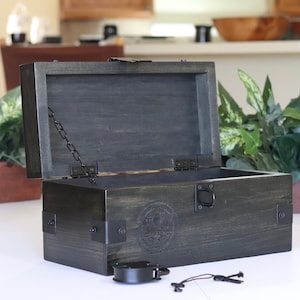 May include: A dark gray wooden box with a hinged lid, black metal hinges, and a black metal chain. The box has a black metal latch and a decorative metal seal on the front. The box is open and the inside is empty.