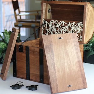 May include: A wooden treasure chest with a hinged lid, black metal accents, and a rope net lining the inside. The chest is open and the lid is propped up. There are two black metal clasps on the lid.