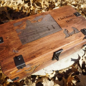 K&ouml;nnte beinhalten: Eine Holzkiste mit einem Scharnierdeckel und Metallverzierungen. Die Kiste ist mit den Worten "Christmas Box" und einem Hausmotiv graviert.