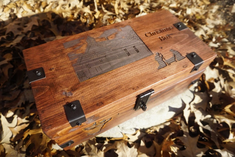 K&ouml;nnte beinhalten: Eine Holzkiste mit einem Scharnierdeckel und Metallverzierungen. Die Kiste ist mit den Worten "Christmas Box" und einem Hausmotiv graviert.