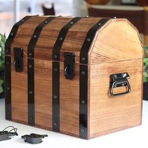 May include: A wooden treasure chest with black metal accents and a hinged lid. The chest has a black metal handle on the front and two black metal clasps on the top. The chest is sitting on a white surface.