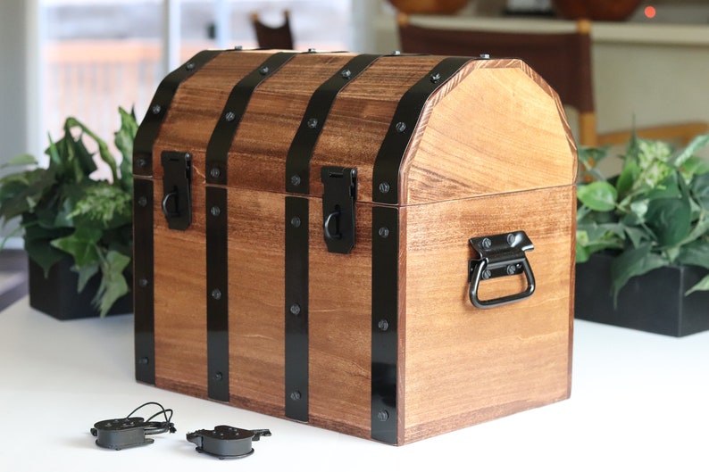 May include: A wooden treasure chest with black metal accents and a hinged lid. The chest has a black metal handle on the front and two black metal clasps on the top. The chest is sitting on a white surface.