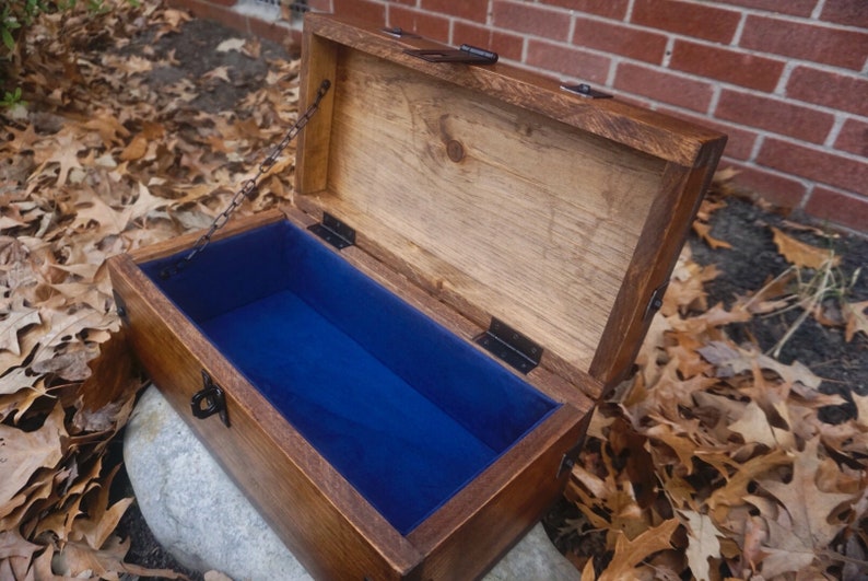 K&ouml;nnte beinhalten: Eine Holzkiste mit einem Scharnierdeckel und einer blauen Samtf&uuml;tterung. Die Kiste hat einen Metallverschluss und eine Kette.