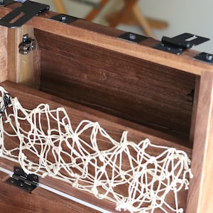 May include: A wooden treasure chest with a hinged lid, black metal accents, and a white rope net lining the inside. The chest has a black metal chain attached to the lid.