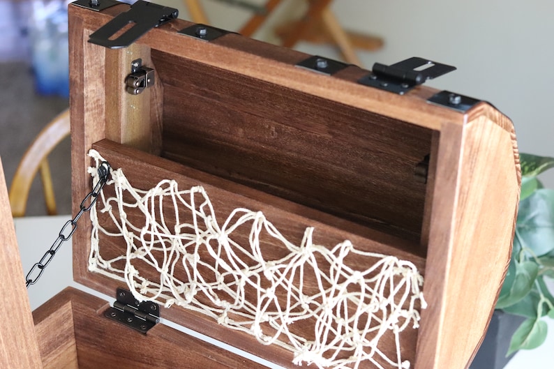 May include: A wooden treasure chest with a hinged lid, black metal accents, and a white rope net lining the inside. The chest has a black metal chain attached to the lid.