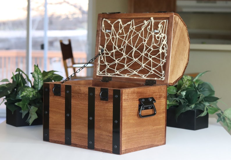 May include: A wooden treasure chest with a hinged lid, black metal accents, and a chain. The lid is open, revealing a white rope net lining the inside.