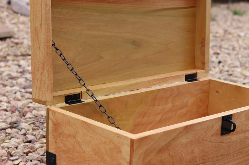 K&ouml;nnte beinhalten: Eine Holzkiste mit einem Scharnierdeckel und einer schwarzen Metallkette. Der Deckel ist ge&ouml;ffnet und das Innere der Kiste ist sichtbar. Die Kiste ist aus hellem Holz gefertigt und hat eine nat&uuml;rliche Oberfl&auml;che.