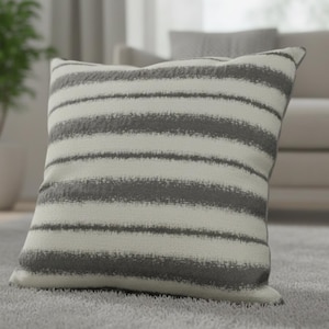 May include: A square cushion featuring a white base with broad, horizontal grey stripes. The cushion is positioned on a grey carpet, suggesting a living room setting. A soft home decor item.