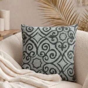 May include: A square decorative pillow with a gray and white swirling pattern rests on a cream-colored armchair. A cream-colored knitted throw blanket is draped over the chair. The background includes a wooden table with candles and a vase.