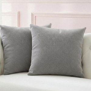 May include: Two square, gray throw pillows rest on a white sofa. The pillows have a solid color and a textured appearance. The sofa has a tufted back and a smooth, white surface. The background is a light pink wall.