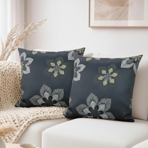 May include: Two square throw pillows with a dark blue background and a floral pattern. The flowers are in shades of white, black, and light green. The pillows are on a white couch with a cream-colored blanket.