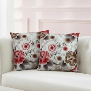 May include: Two square throw pillows with a floral pattern. The pillows feature large, stylized flowers in shades of red, pink, and brown against a cream-colored background. The pillows are placed on a white sofa.