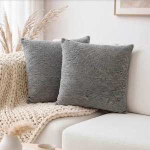 May include: Two square, gray throw pillows with a textured surface rest on a white couch. A cream-colored, chunky knit blanket is draped over the couch. Dried ornamental grass and a small white vase are visible.