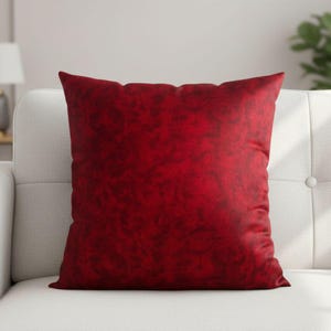 May include: A square, red decorative pillow with a textured surface. The pillow is placed on a white sofa. The pillow's color is a deep, rich red, adding a pop of color to the neutral background.