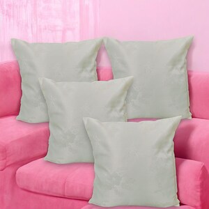 May include: Four white throw pillows with a subtle floral pattern are arranged on a pink couch. The pillows are made of a soft, textured fabric.