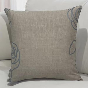 以下が含まれることがあります： ニュートラルなベージュの生地を使用した正方形の装飾用クッション。角には、落ち着いたグレーブルーの色合いの繊細な花の模様があります。白いソファの上に置かれており、インテリアのテーマを示唆しています。