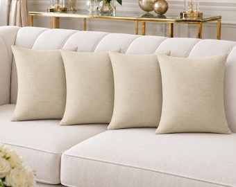 May include: Four square scatter cushions in a solid light beige colour with a visible woven linen-style texture. The cushions are arranged in a row along the back of a matching light beige upholstered sofa, featuring a clean and simple design.