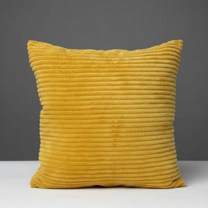 May include: A square, mustard-yellow corduroy pillow. The pillow has a textured, ribbed surface and is placed on a white surface against a gray background. The pillow is a solid color and has a soft, inviting appearance.