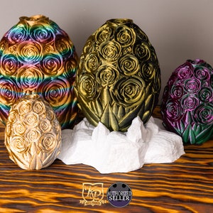 May include: Four decorative dragon eggs with a rose pattern. The eggs are in various colors, including gold, rainbow, and purple. The eggs are sitting on a white rock-like base.