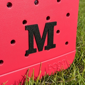 May include: A bright red cooler bag with a black letter "M" on the front. The cooler bag is made by the company "bogg" and is sitting on green grass.
