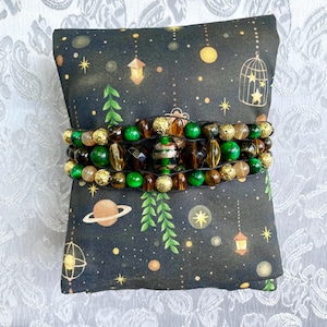 May include: A beaded bracelet with green, brown, and gold beads is displayed on a black pillow with a celestial pattern. The bracelet features various bead shapes and sizes, including round, faceted, and cylindrical beads. The pillow has a print of planets, stars, and hanging lanterns.