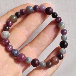 以下が含まれることがあります： 様々な色の宝石をあしらったビーズブレスレット。紫、グレー、茶色の丸いビーズが含まれています。白い背景に、手でブレスレットを持っています。