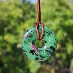 Può includere: Un ciondolo in pietra verde e nera con un foro centrale, infilato su un cordino di cuoio marrone. La pietra presenta un motivo maculato con sfumature rosa. Lo sfondo è una vista sfocata di fogliame verde.