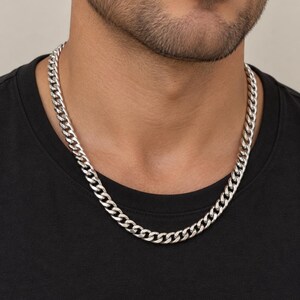 Puede incluir: Un collar de cadena tipo barbada en tono plateado brillante con eslabones planos y ovalados entrelazados. El collar descansa sobre una camiseta negra lisa, mostrando un diseño clásico y uniforme de eslabones metálicos alrededor del cuello.
