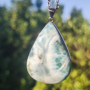 Puede incluir: Un collar con colgante en forma de lágrima con una piedra de larimar pulida. La piedra muestra una mezcla de blanco, azul claro y turquesa, con una cadena plateada. El fondo es un follaje verde borroso.