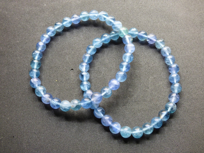 May include: Two blue gemstone bracelets made of round beads. The bracelets are intertwined and are on a gray background.