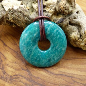 Pode incluir: Um pingente de pedra aventurina verde-azulada em forma de rosquinha, com um cord&atilde;o de couro marrom.