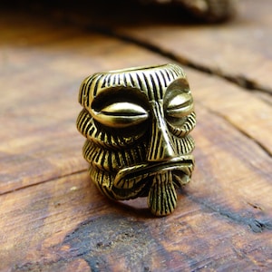 Puede incluir: Una pequeña cuenta con forma de máscara tiki de color latón, con un diseño tallado detallado. La cuenta presenta una cara estilizada con los ojos cerrados, una nariz prominente y una boca hacia abajo. La cuenta es cilíndrica y tiene un agujero en el centro.