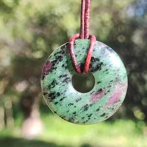 Può includere: Un ciondolo rotondo in zoisite verde con inclusioni di rubino, sospeso da un cordino in pelle marrone. La pietra ha un foro centrale ed è macchiata di nero e rosa. Lo sfondo è verde sfocato.