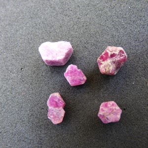Könnte beinhalten: Fünf grob geschnittene Rubin-Edelsteine in verschiedenen Formen und Größen. Die Edelsteine weisen eine Reihe von rosa und roten Farbtönen auf, wobei einige ein gesprenkeltes Aussehen haben. Sie liegen auf einem dunkelgrauen, strukturierten Hintergrund.