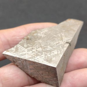 May include: A triangular-shaped metallic meteorite fragment with a polished surface displaying a distinct Widmanstätten pattern of intersecting crystalline lines in shades of silver, grey, and muted brown, showing a sharp geometric edge.