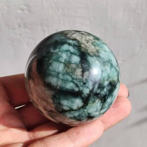 Pode incluir: Uma esfera de esmeralda verde com um padrão de redemoinhos de veios verde escuro e branco. A esfera é lisa e polida.