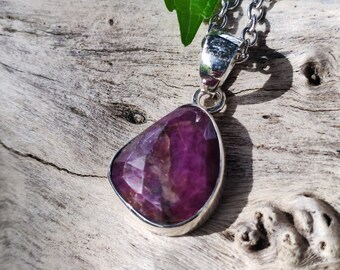 Puede incluir: Un collar con colgante de plata con una gran piedra preciosa rosa oscuro facetada en forma de lágrima. El colgante está engastado en un bisel de plata y cuelga de una delicada cadena de plata. Una hoja verde es visible en el fondo.