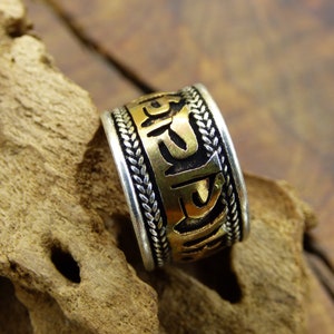 Puede incluir: Un anillo de plata y oro con escritura grabada. El anillo tiene una banda ancha con una sección central dorada con letras oscuras. Los bordes son plateados con un diseño trenzado. El anillo se exhibe sobre un trozo de madera.